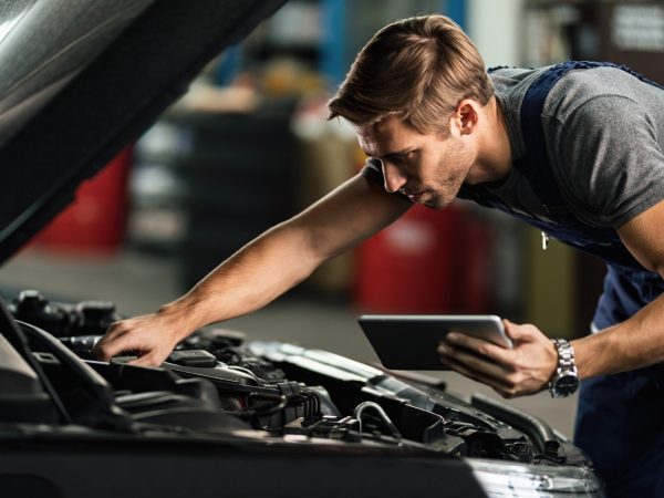 Young auto mechanic using digital tablet while repairing car engine in a workshop.